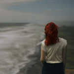 Redhead woman walking by ocean meditating on Greatest Overcomer